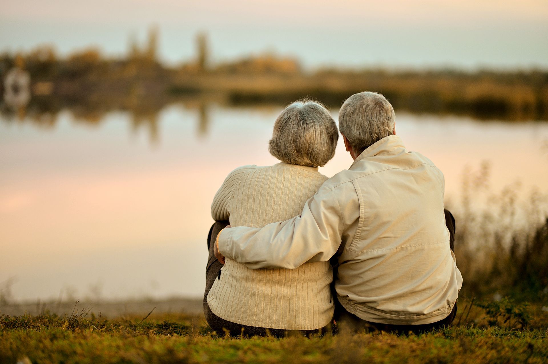 Couple with gray hair sits with arms around each other, looking at calm water and distant landscape at sunset.