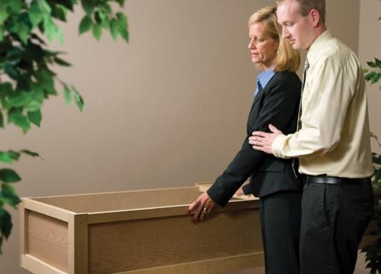 Woman and man at a funeral, looking at an open casket in a beige room. The man has his hand on the woman's arm.