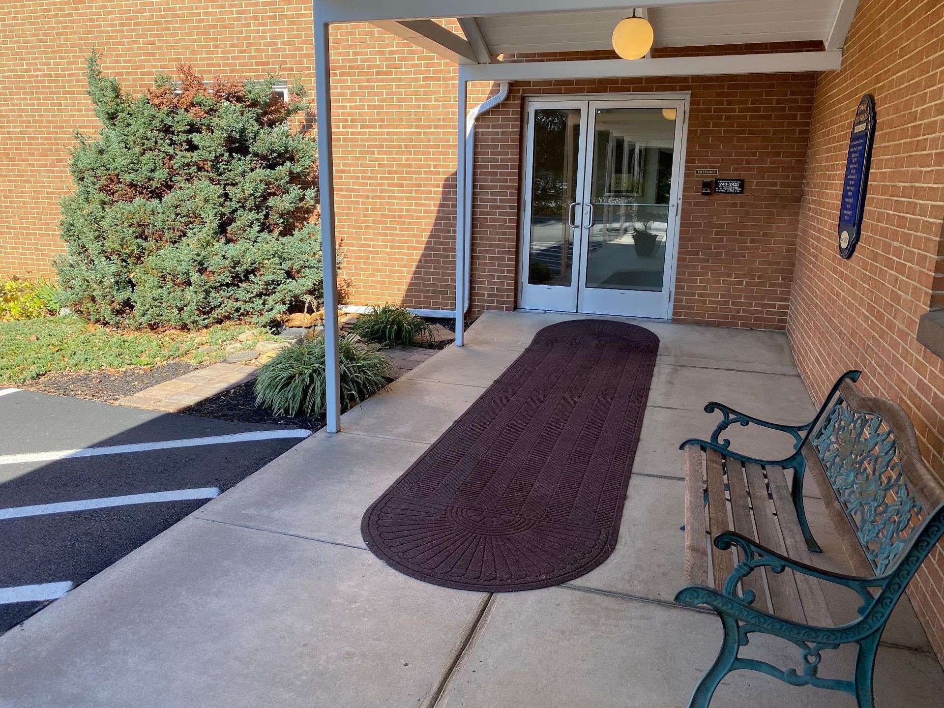 Entrance to a brick building with a covered walkway, brown rug, bench, and double doors.
