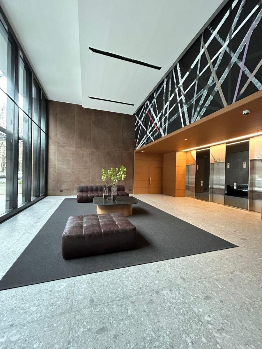 Vestíbulo de edificio moderno con alfombra gris oscuro, asientos marrones y grandes ventanales.