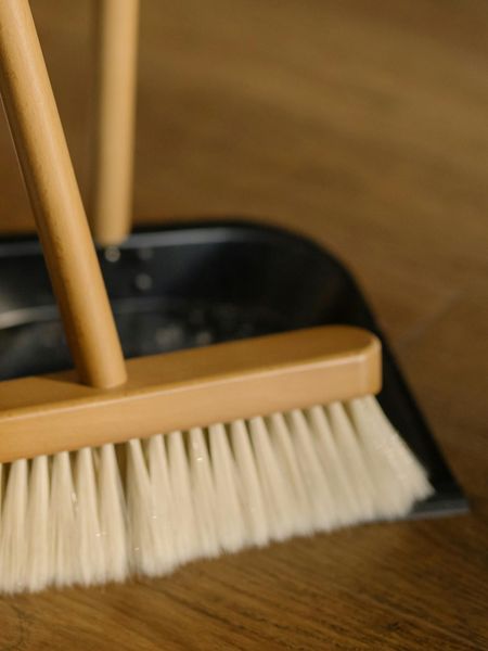 Broom with light brown handle and light bristles in a black dustpan on a wooden floor.