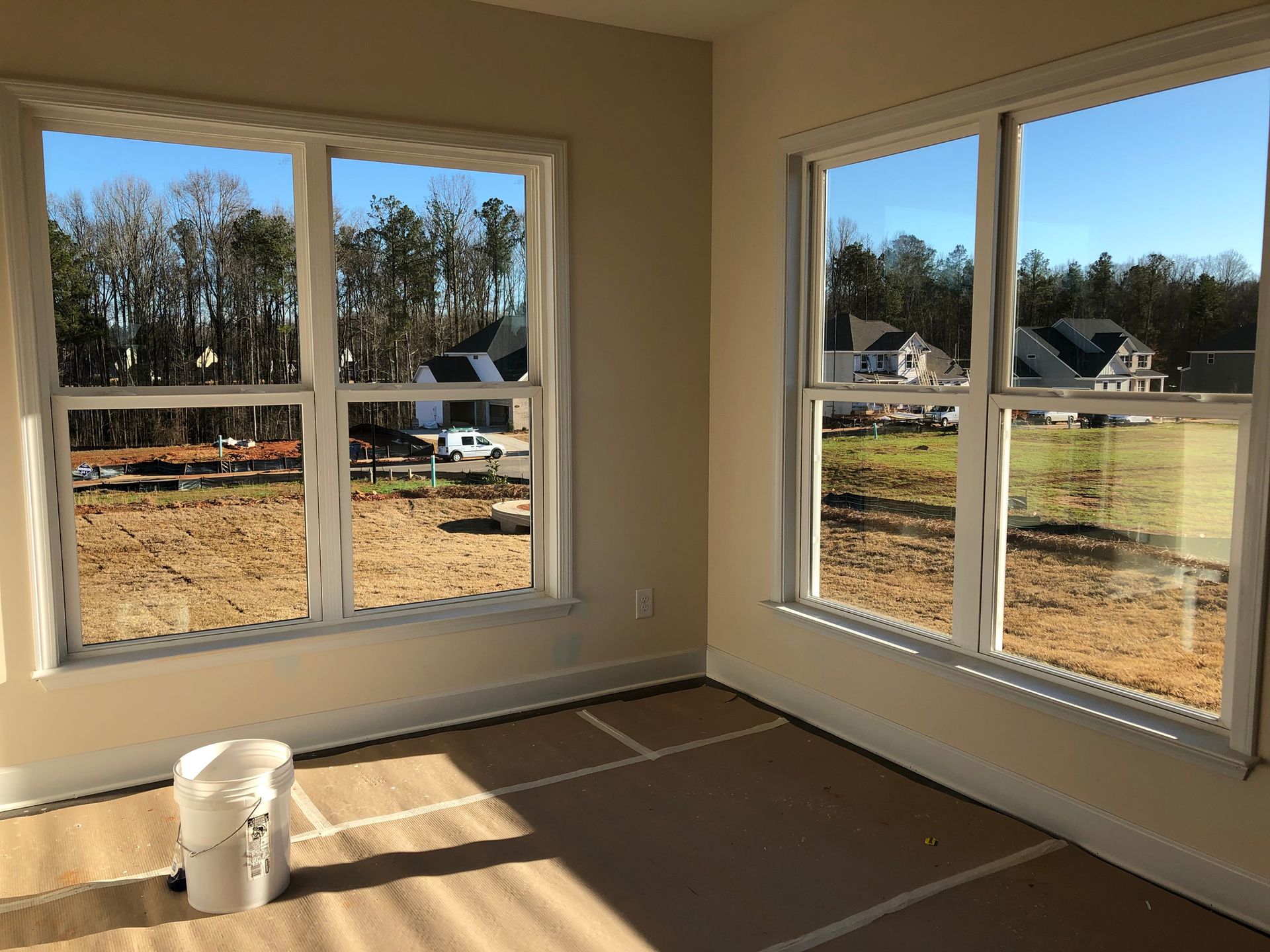 Interior view of a room with two large windows overlooking a construction site and residential area. The room is unfinished with a paint bucket on the floor.
