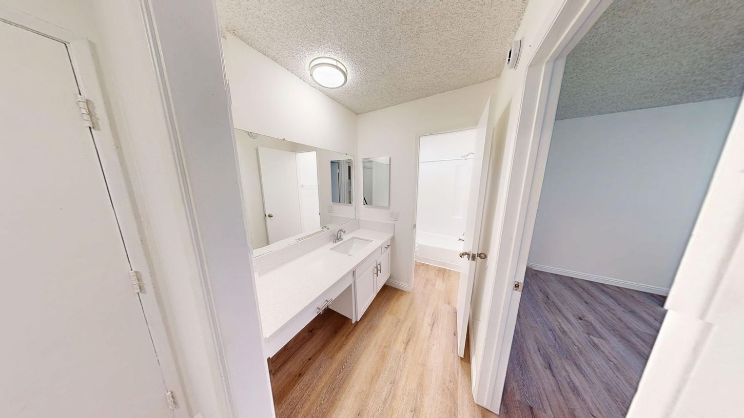 Bathroom with white walls, long vanity, and adjacent room with wood-look flooring.