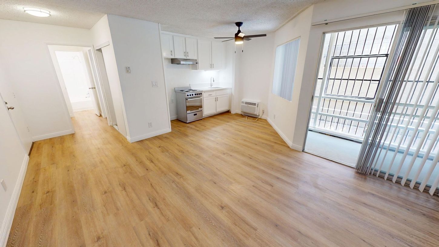 Interior view of a small, bright apartment with wood flooring, white walls, and a small kitchen.