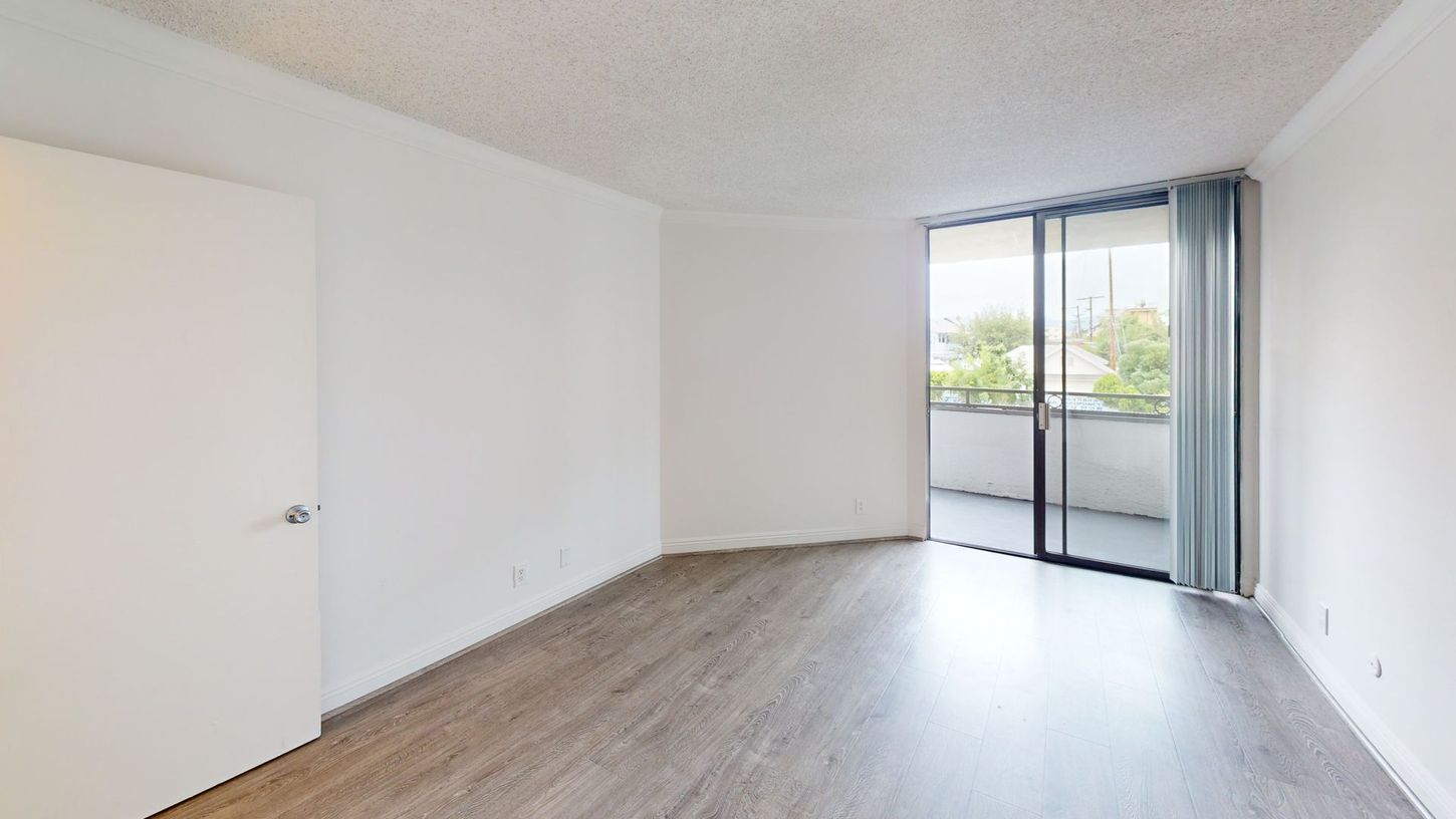 Empty room with wooden floor, white walls, balcony, and closed sliding glass door.
