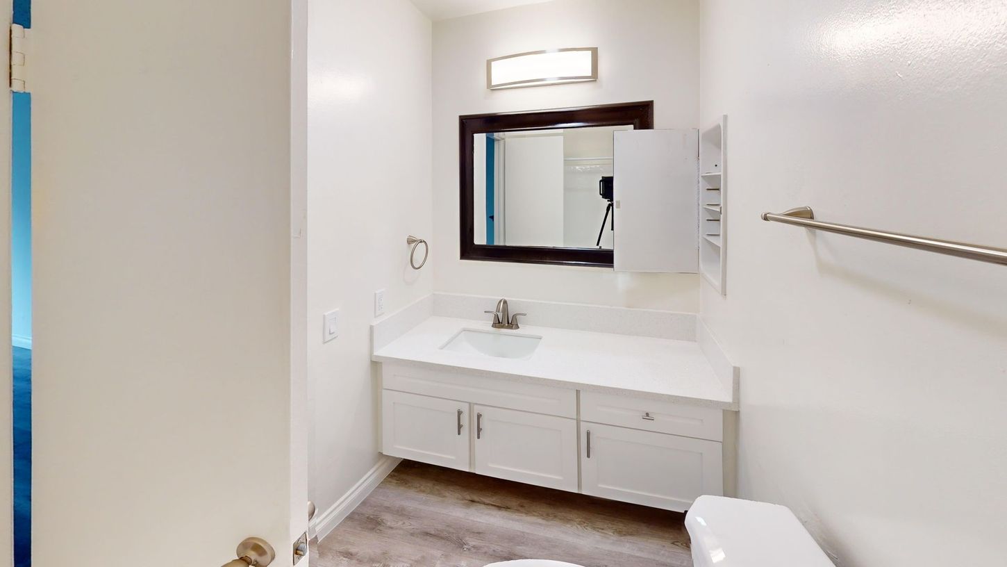 White bathroom with vanity, mirror, and toilet. Light fixtures and towel rack are visible.