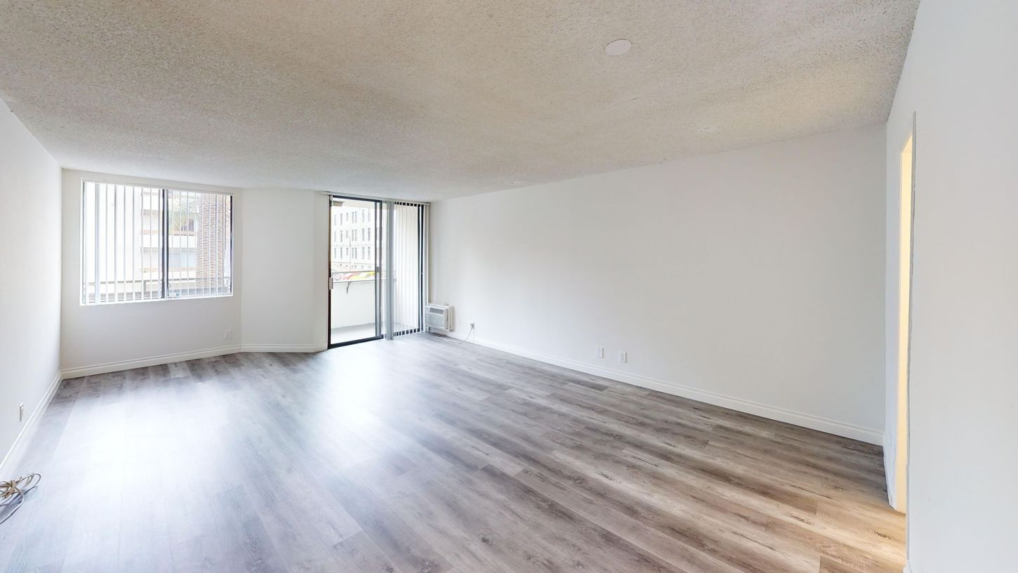 Empty, white-walled room with gray wood-look flooring, large windows, and a sliding door leading to a balcony.