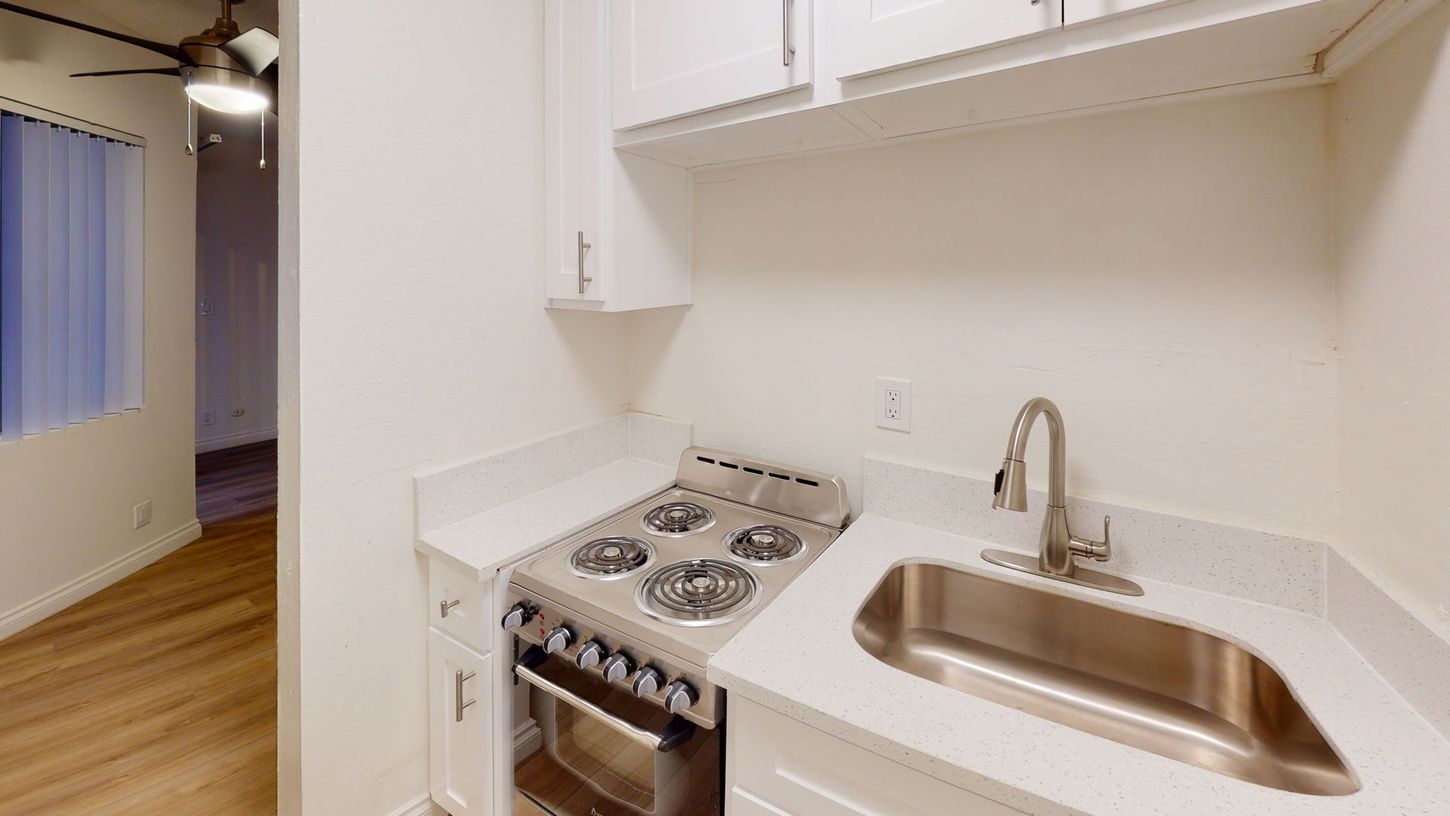 Small kitchen with a stove, sink, and cabinets. Bright white walls and countertops, wood flooring in the background.