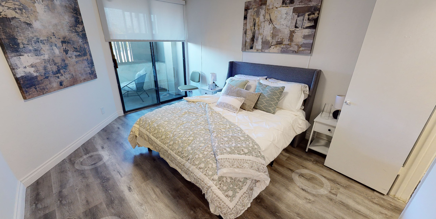 Bedroom with bed, art, and balcony. Light gray wood flooring, white walls, and bright sunlight.