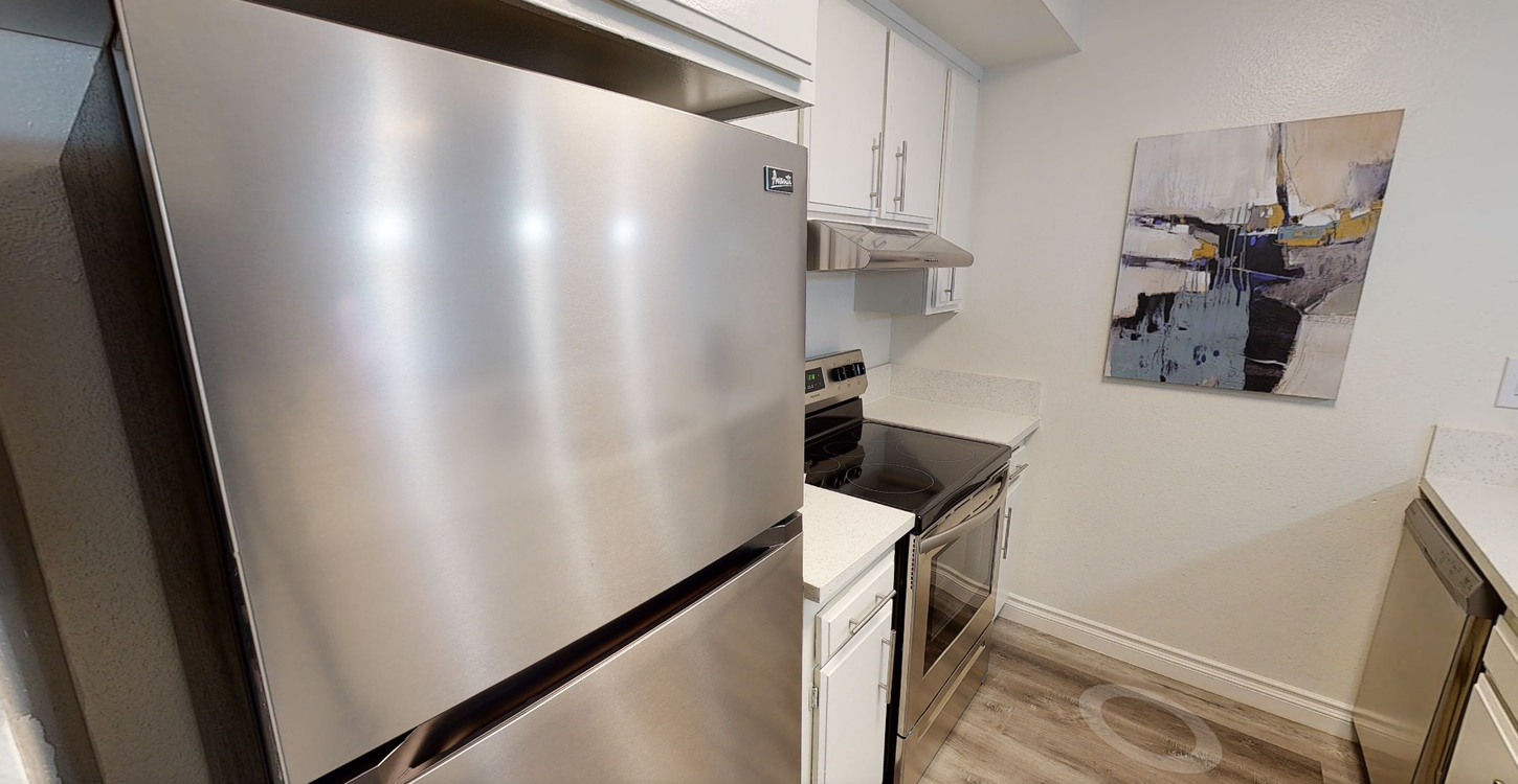 Stainless steel refrigerator in a small kitchen with white cabinets, stovetop, and abstract art on the wall.