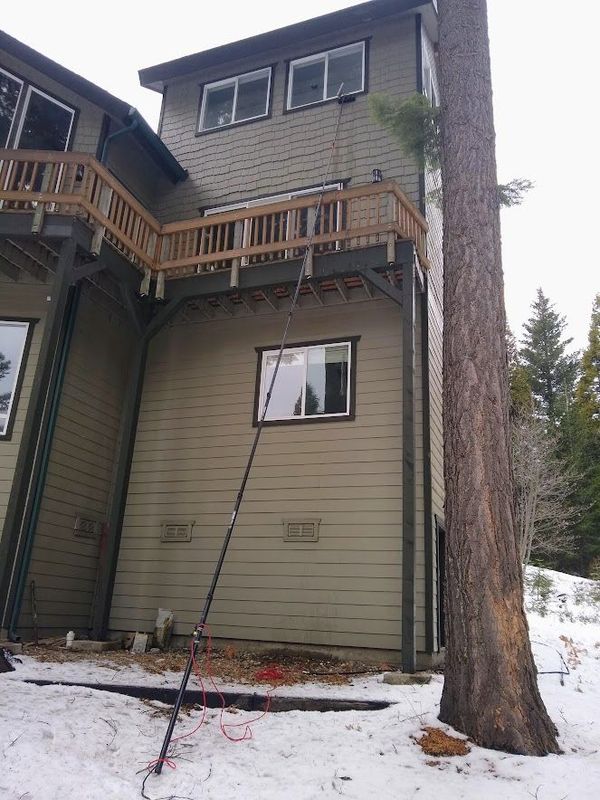 A long pole is extended to retrieve a cord hanging from the wooden deck of a multi-story house surrounded by snow and trees.