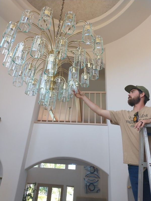 A person on a ladder reaching up to adjust a large, tiered, brass-and-glass chandelier in a high-ceilinged room.