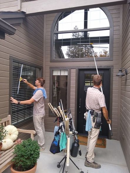 Two professional window cleaners clean a home's exterior windows using long-reach squeegees and a tool-filled cart.