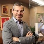 Man in glasses and blazer smiles, arms crossed, in office setting.