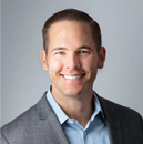 Man smiling, wearing a blue shirt and gray blazer against a gray background.