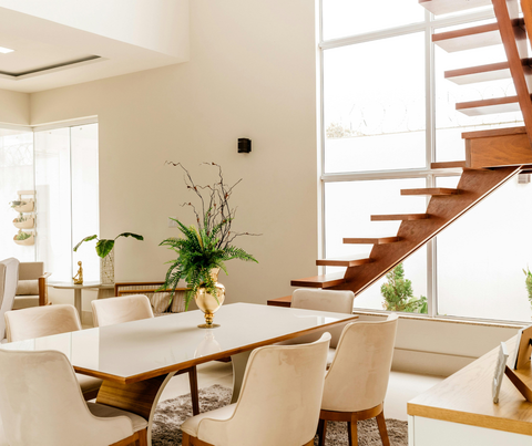 Bright modern dining area with wooden staircase, white chairs, and potted plants near large windows