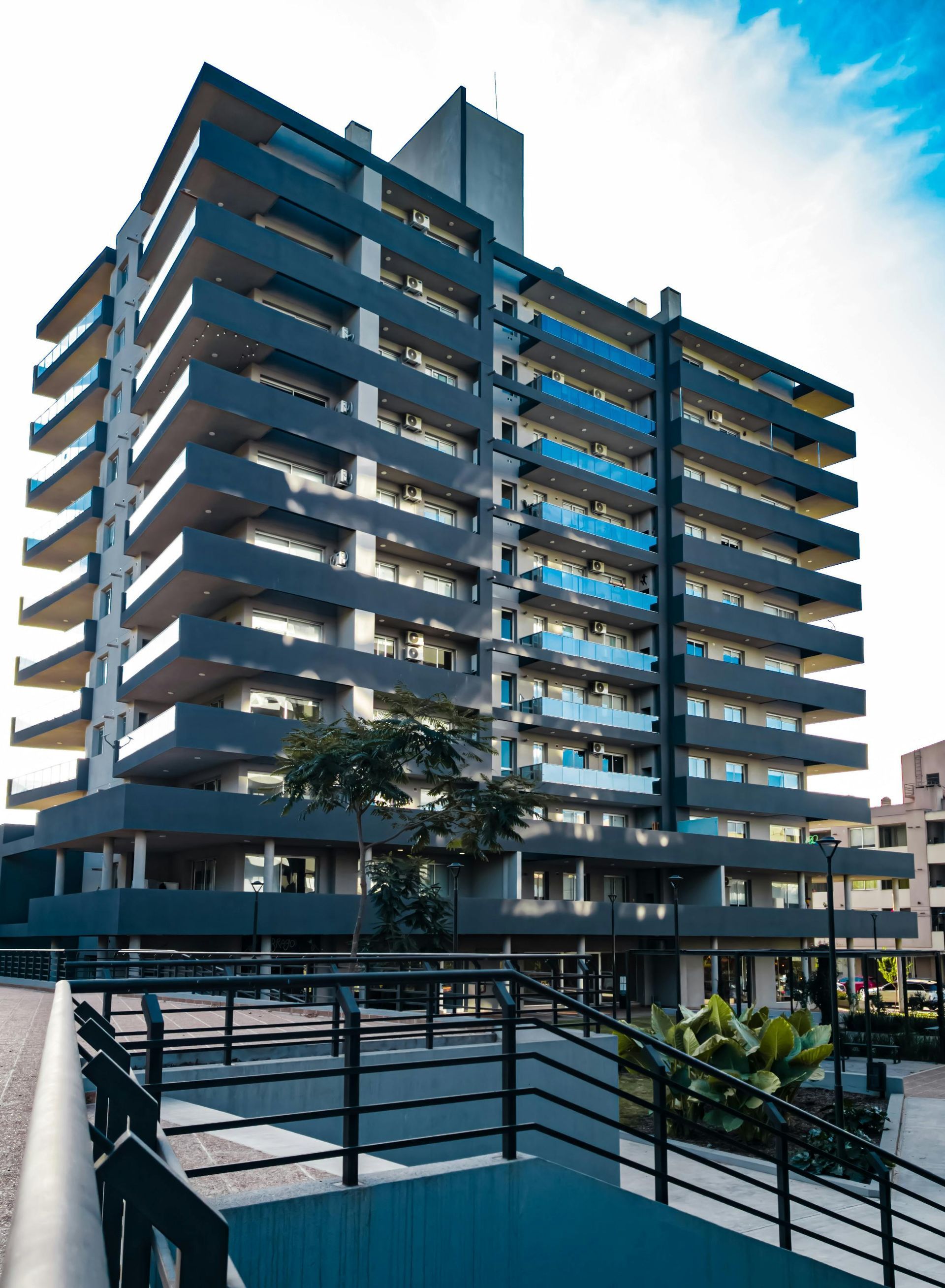 A modern multi-story apartment building with dark blue balconies and a tiered exterior, viewed from a pedestrian ramp.