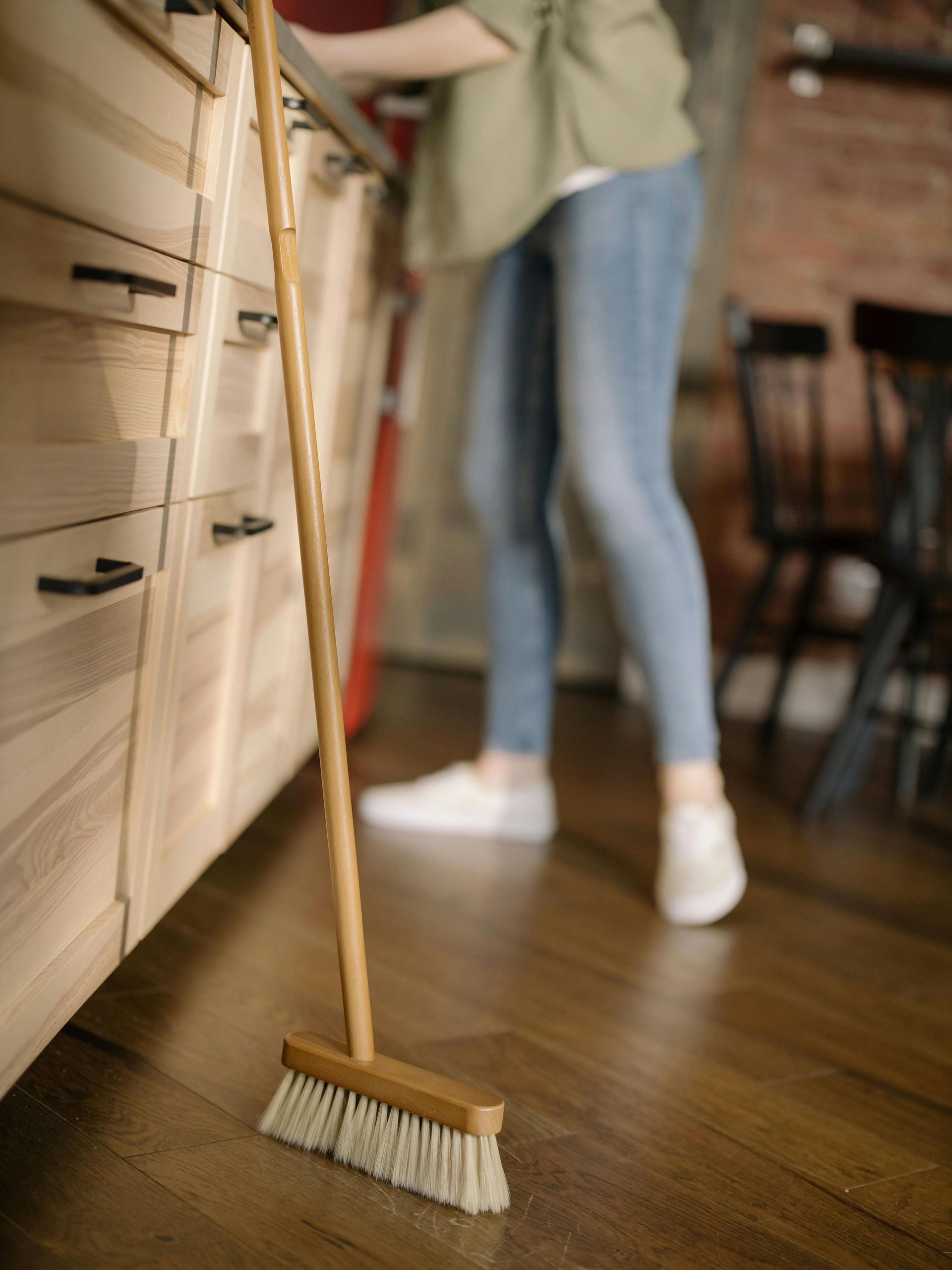 A wooden broom stands upright on a dark wooden floor in a kitchen, with a person in jeans standing blurred in the background.