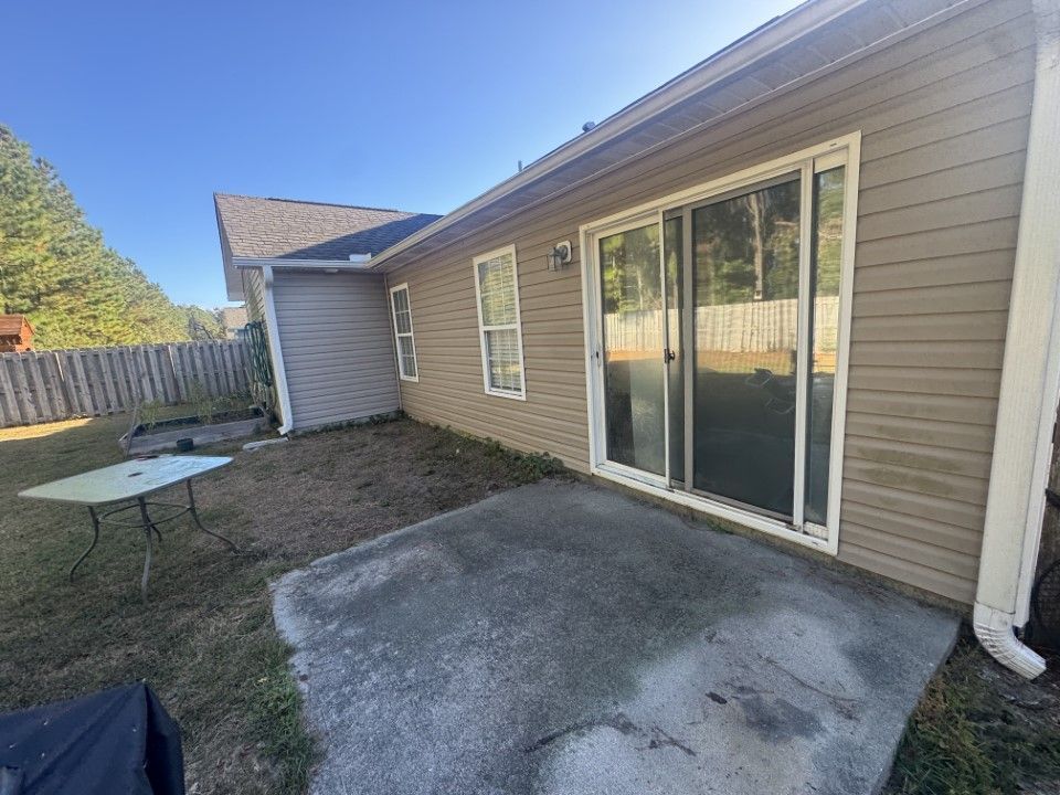 Backyard with concrete patio, sliding glass door, tan siding, and a fence. A table sits on the grass.