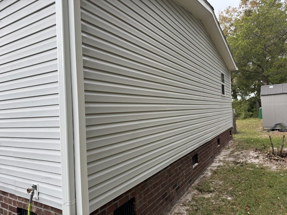 Side of a house with light gray siding and brick foundation. A small window and a shed are visible.