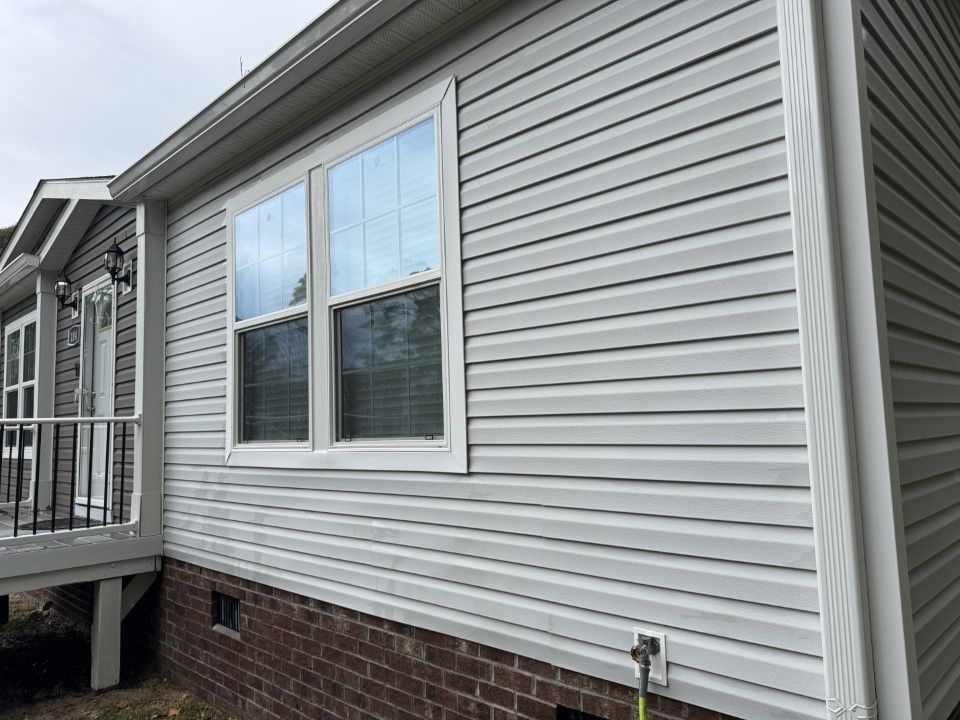 Exterior of a house with gray vinyl siding, white window trim, and a brick foundation.