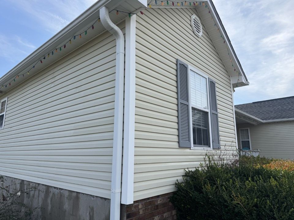Beige vinyl-sided house with white trim, window, and gray shutters.