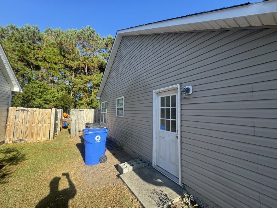 Gray house exterior with a white door, blue trash can, and a wooden fence. Sunny day.