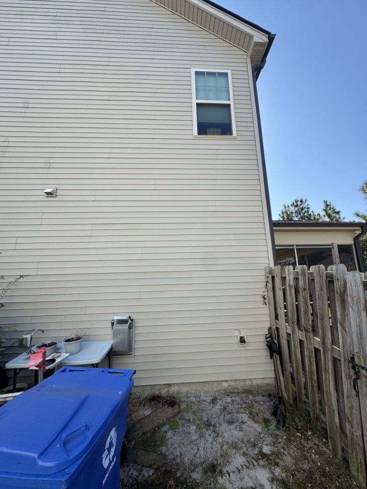 Side of a beige house with a window and a blue recycling bin in front of it, next to a wooden fence.