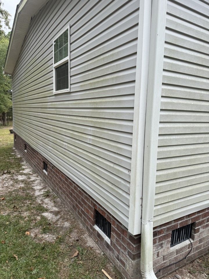 Side view of a house with light gray siding, a brick foundation, and a white downspout; appears dirty.