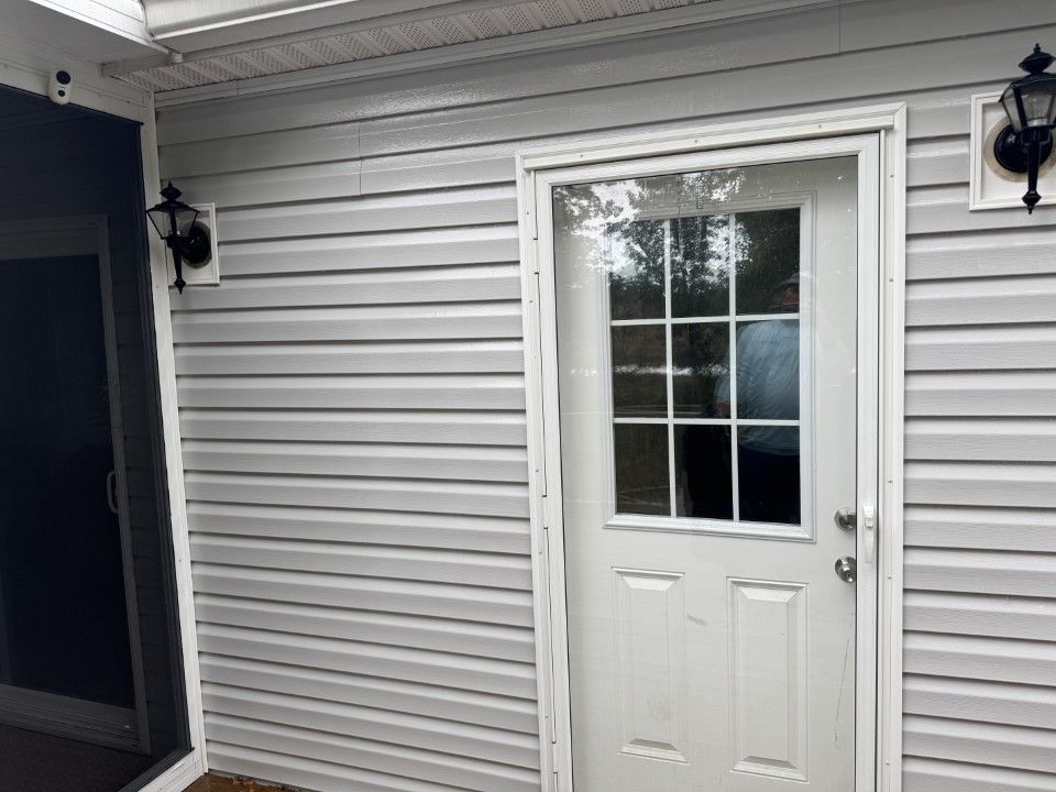 Exterior door with glass panels and sidelights, flanked by sconces, set in a light gray-sided wall.
