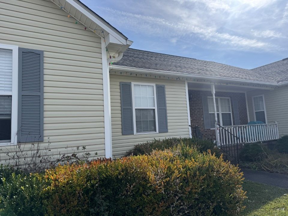 Beige vinyl-sided house with grey shutters and porch; bushes in front.