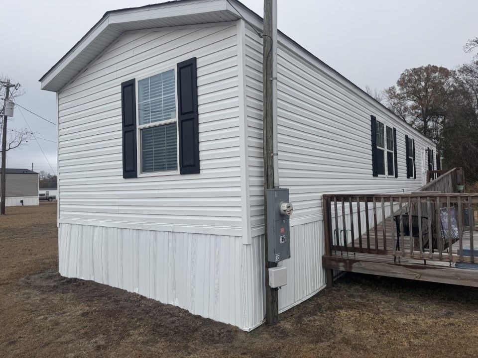 White mobile home with black shutters, gray electrical box, and small wooden deck.