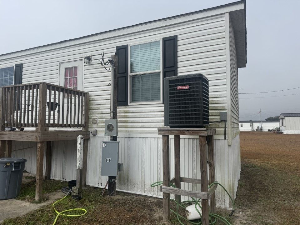 Mobile home exterior with a Goodman AC unit on a wooden stand. Gray siding, black shutters.