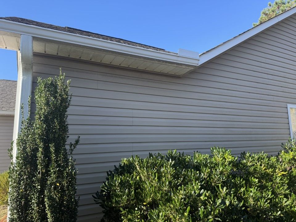 Tan vinyl siding on a house with white trim, gutters, and bushes in front. Clear blue sky.