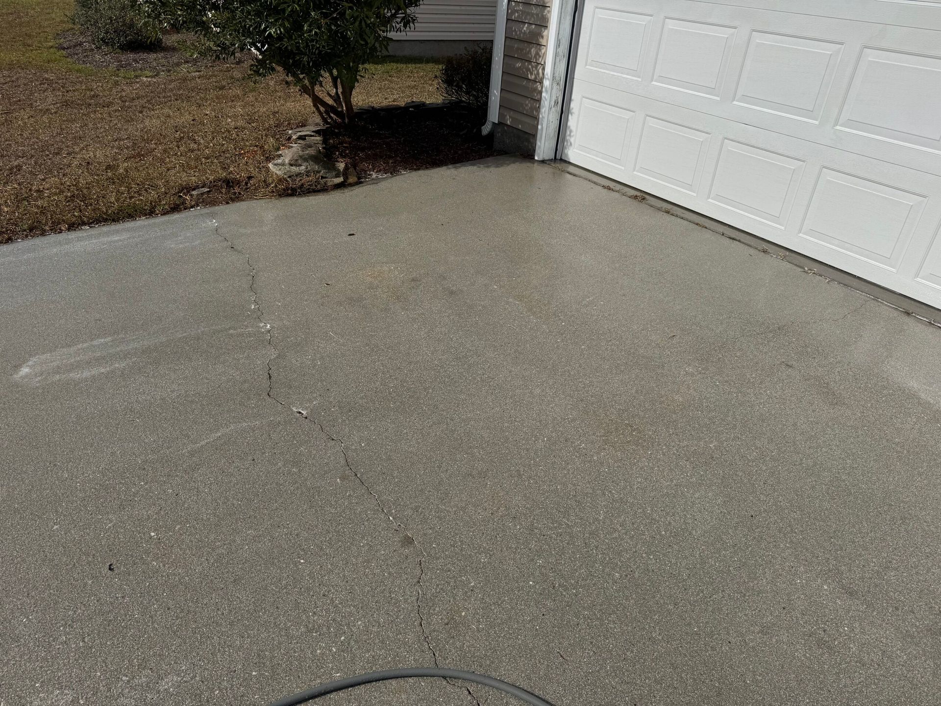 Concrete driveway next to a white garage door, with a small bush and brown grass.