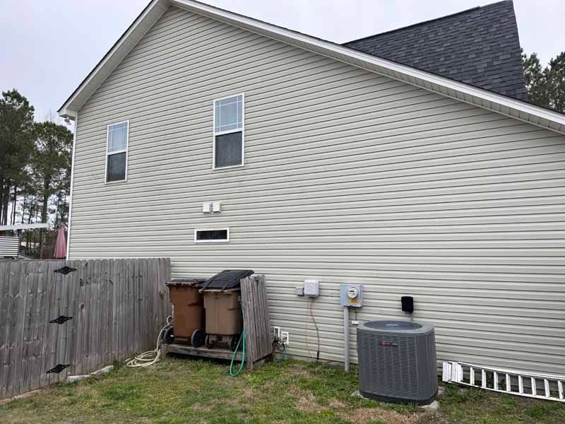 Side of a house with beige siding, two windows, and an air conditioning unit. A wooden fence is in the foreground.