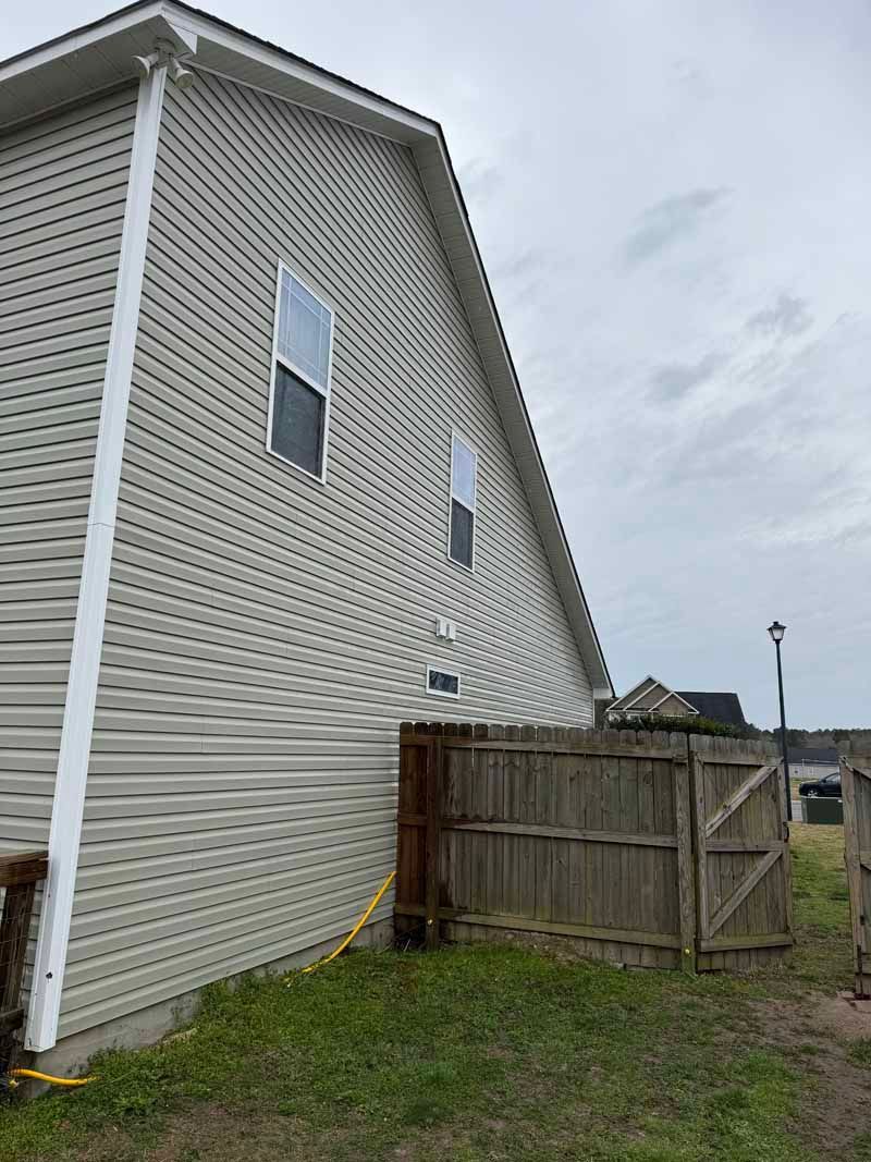 Side of a light beige house with white trim, two windows, next to a wooden fence and gate.