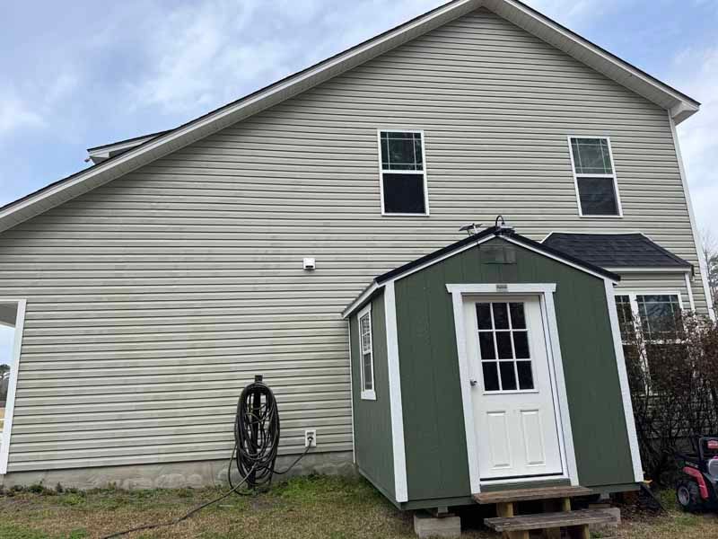 Side view of a two-story beige house with a green shed attached. A black hose is mounted on the wall.