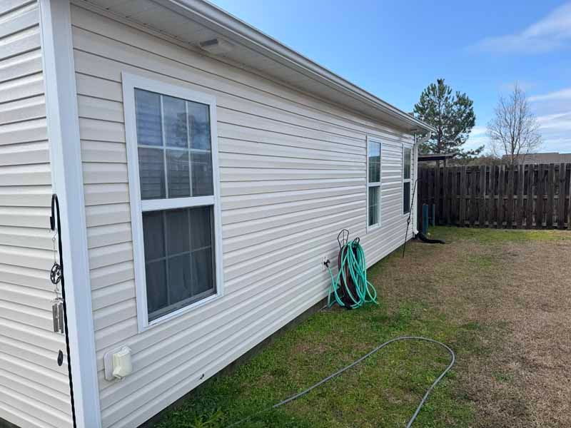Beige siding on a house with white-framed windows, green hose, and a brown wooden fence on a sunny day.
