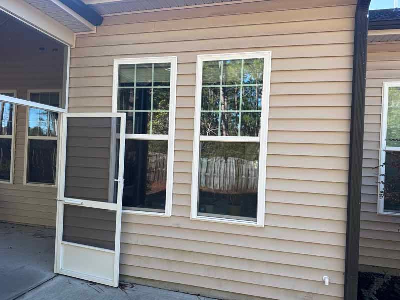 Exterior view of a beige-sided house with two white-framed windows; a screen door leans against the wall.