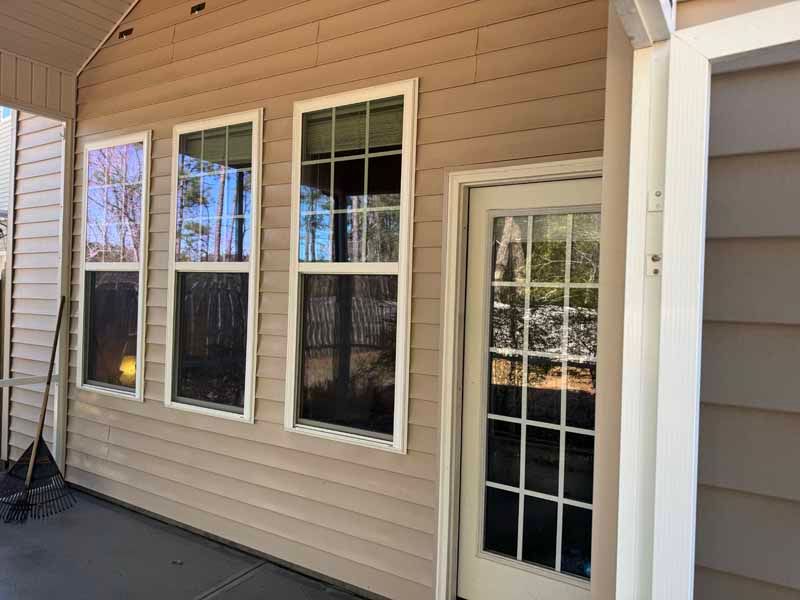 Exterior view of a house with three windows and a door. Tan siding, white trim, reflecting trees.