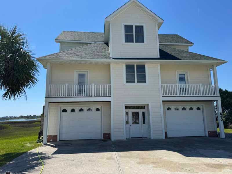 Multi-story beige house with white trim, balconies, and garage doors; a palm tree and blue sky visible.