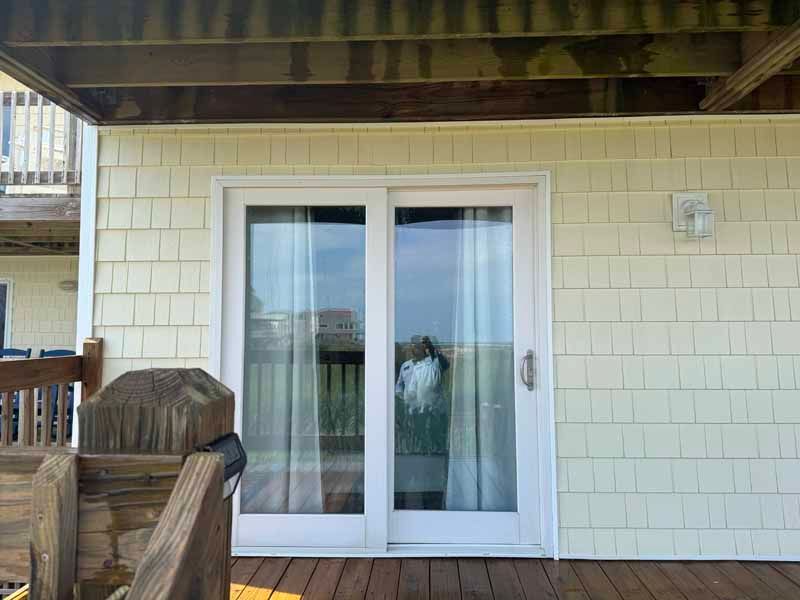 White sliding glass doors on a deck, beneath a wood overhang, surrounded by yellow shingle siding.