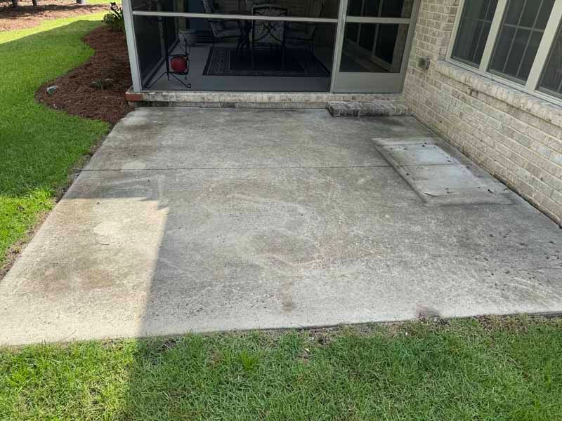 Concrete patio with discoloration, adjacent to a screened porch and brick wall, surrounded by grass.