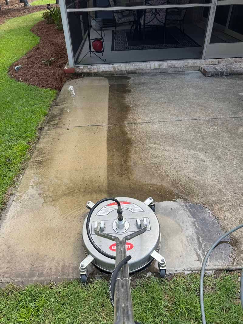 Pressure washer cleaning a concrete patio; half cleaned, half dirty; green grass, screened porch in background.