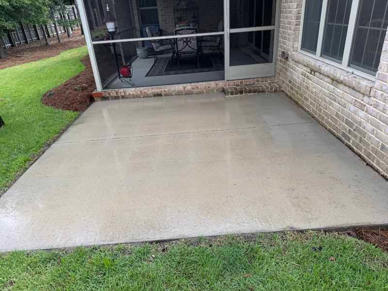 Concrete patio next to a brick wall and screened-in porch, surrounded by grass and mulch.