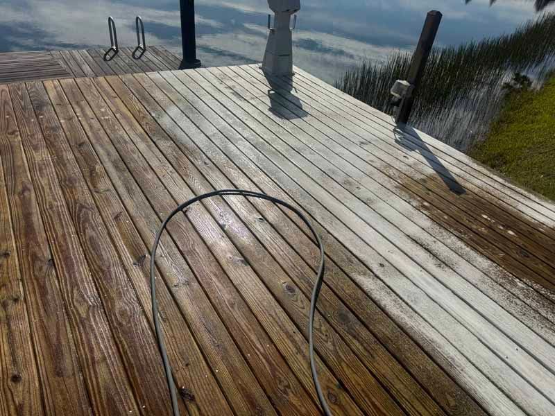 Wooden deck being cleaned with a hose, showing contrast between cleaned and uncleaned sections, near water.