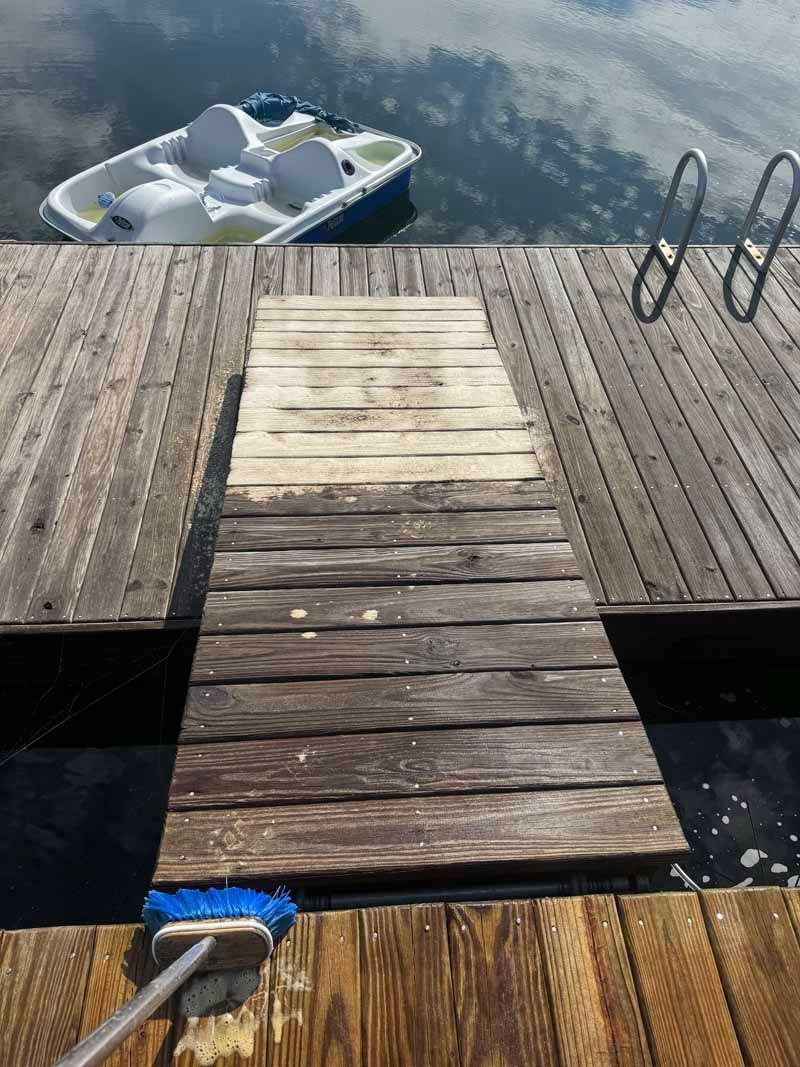 A person is scrubbing a wooden dock with a blue brush. A small boat floats in the water nearby.
