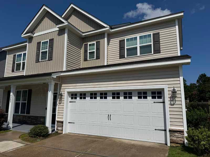 Two-story house with a white garage door, tan siding, brown shutters, and blue sky.