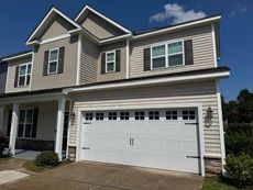 Two-story house with a white garage door, tan siding, brown shutters, and blue sky.